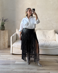 Woman taking a mirror selfie wearing a white shirt and black fringe skirt in a living room.