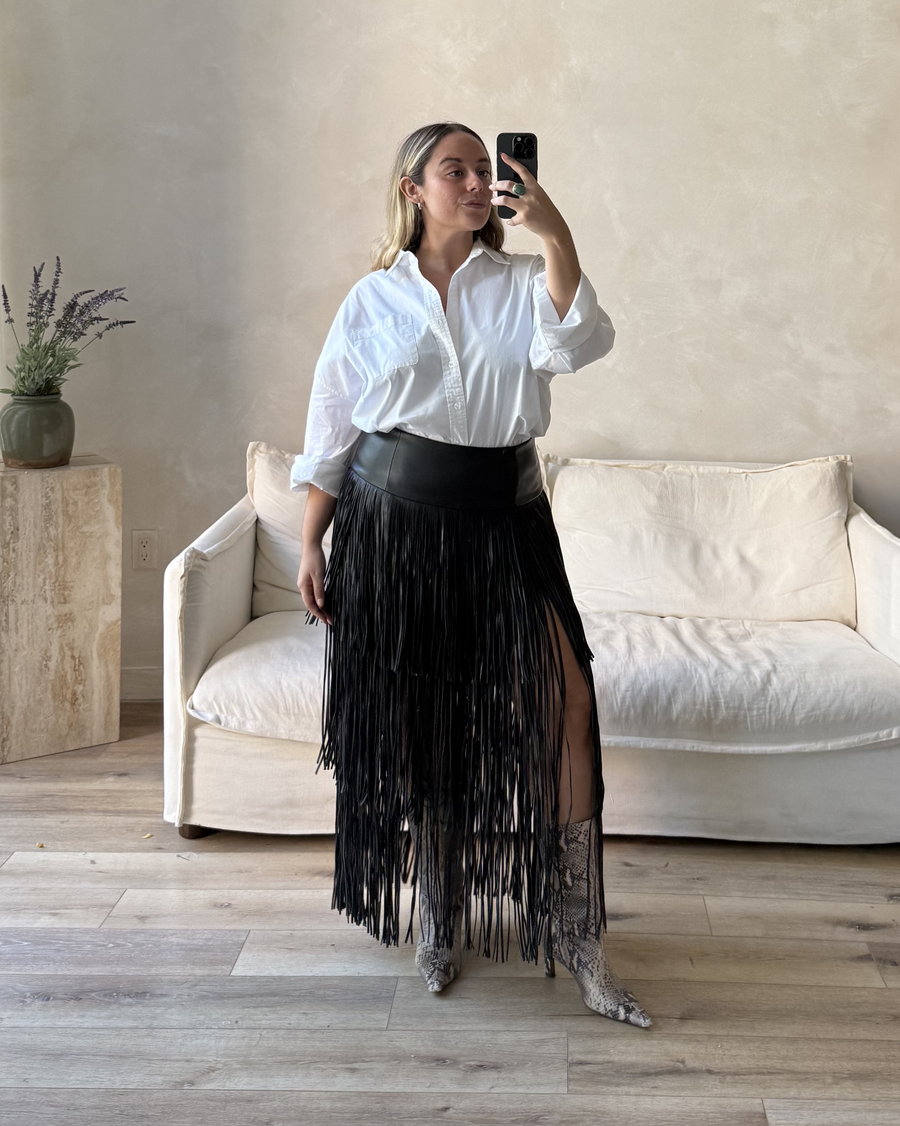 Woman taking a mirror selfie wearing a white shirt and black fringe skirt in a living room.