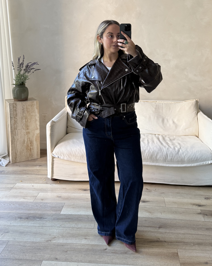 Person wearing a dark brown leather jacket and blue jeans taking a mirror selfie in a room with a white couch and wooden side table.